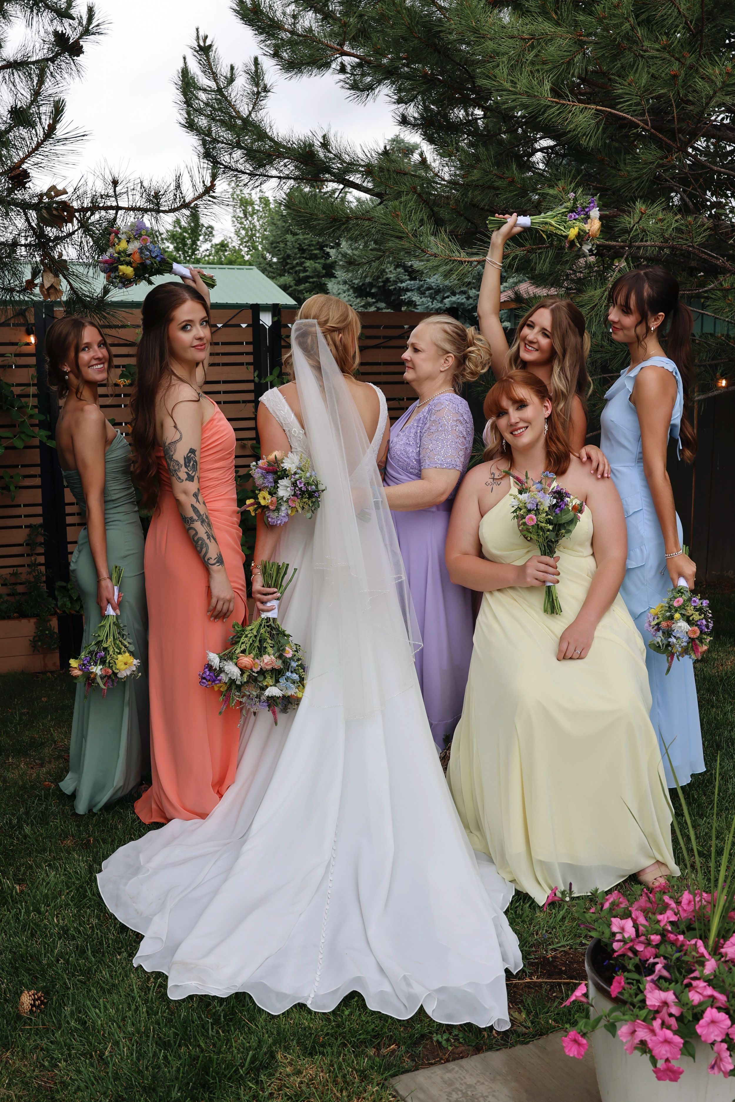 Bride in white gown with veil poses with bridesmaids in colorful pastel dresses holding floral bouquets outdoors.