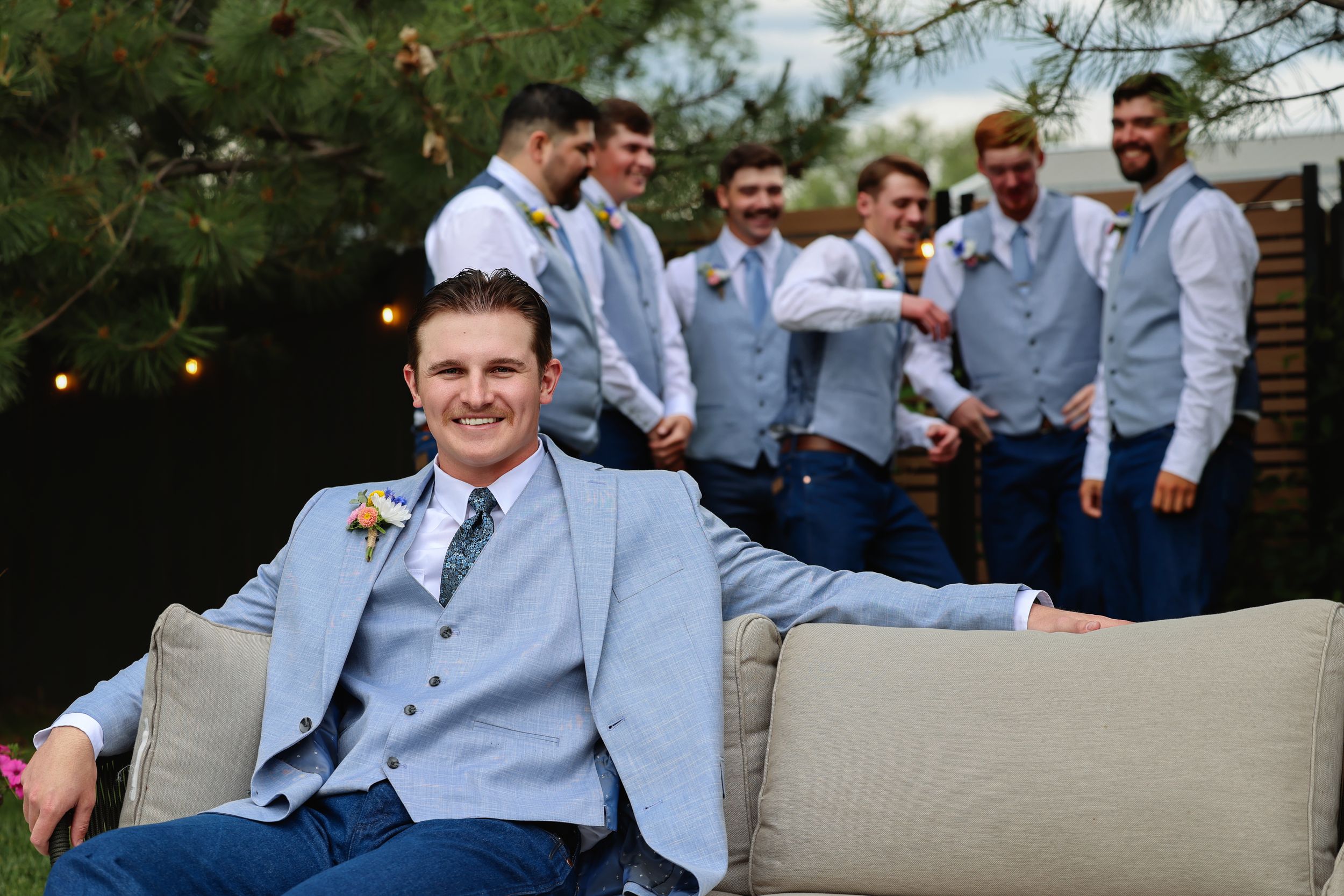 Groom in light blue suit relaxes on outdoor sofa with groomsmen laughing in background at wedding.