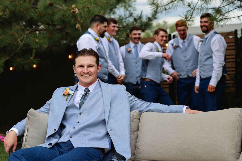 Groom in light blue suit relaxes on outdoor sofa with groomsmen laughing in background at wedding.