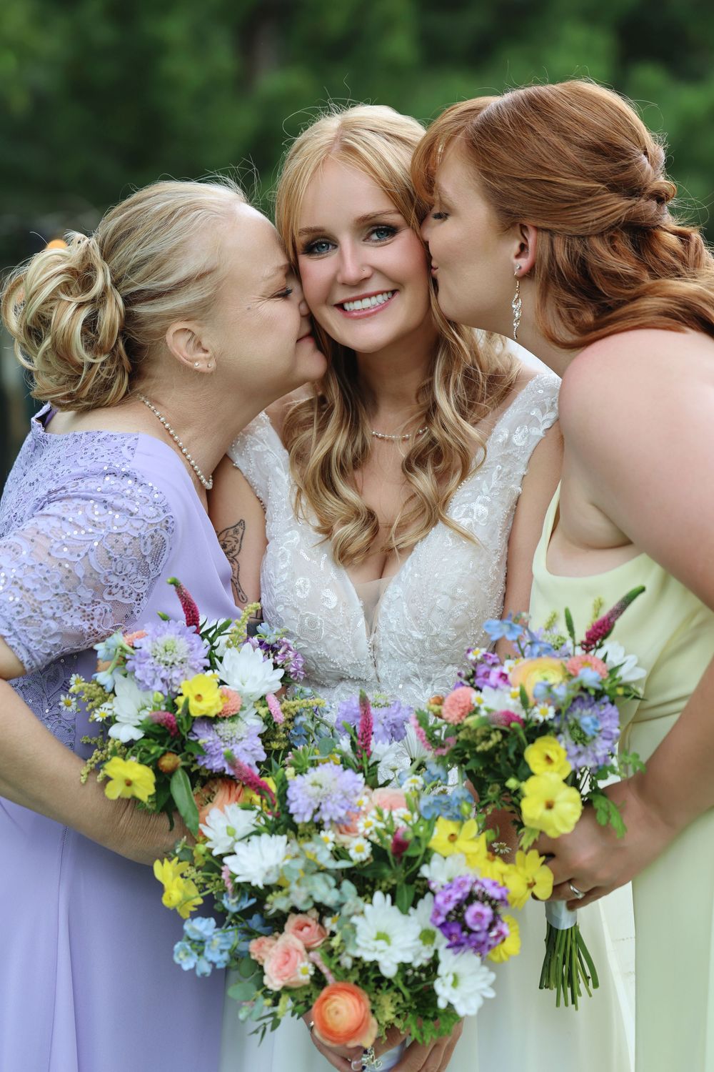 Bride smiling as two bridesmaids kiss her cheeks, holding colorful spring bouquets at an outdoor wedding.