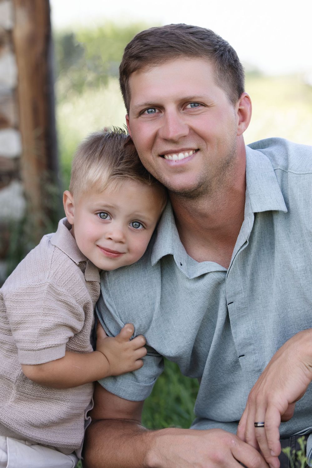 A smiling father and young blonde son pose cheek-to-cheek outdoors in a warm, candid family portrait.