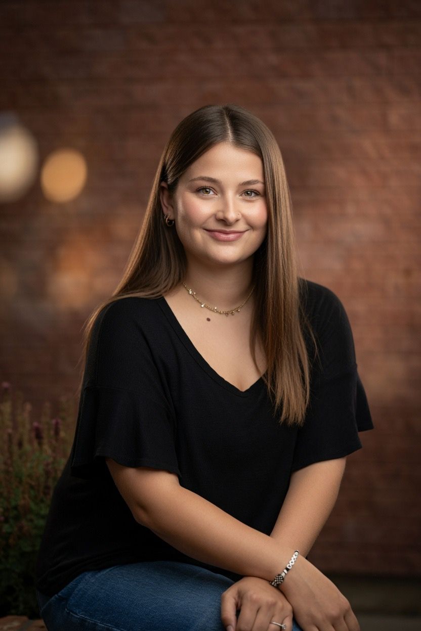 Young woman in black top and jeans smiling, seated against a rustic brick wall backdrop.