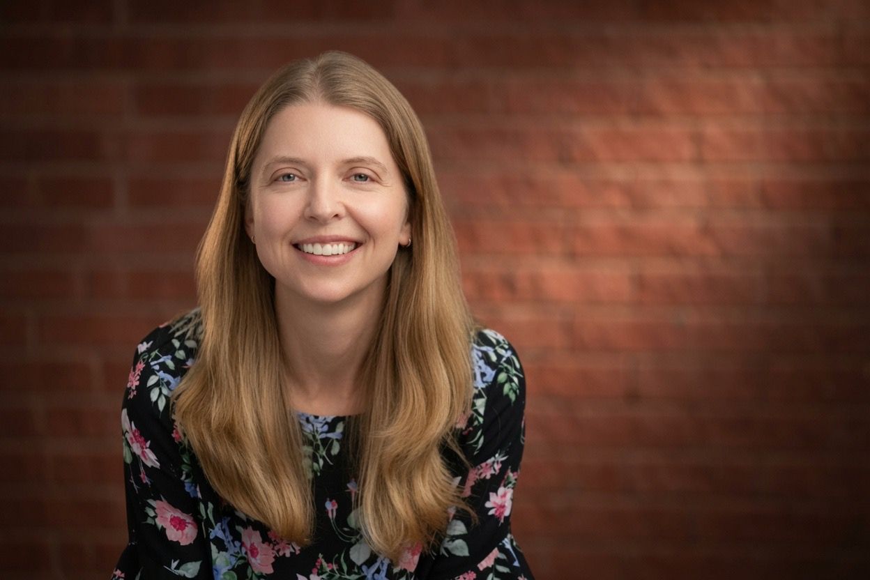 Smiling woman with long blonde hair wearing a floral top, posing against a warm red brick wall background.