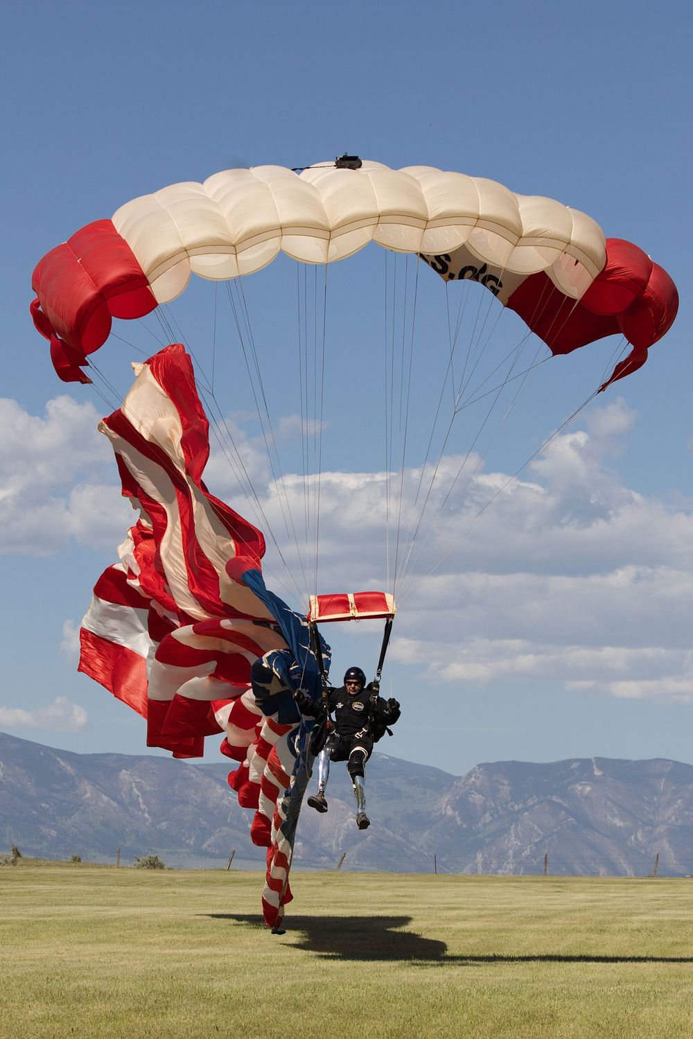 Skydiver landing with American flag under red and white parachute on green field with mountains in background.