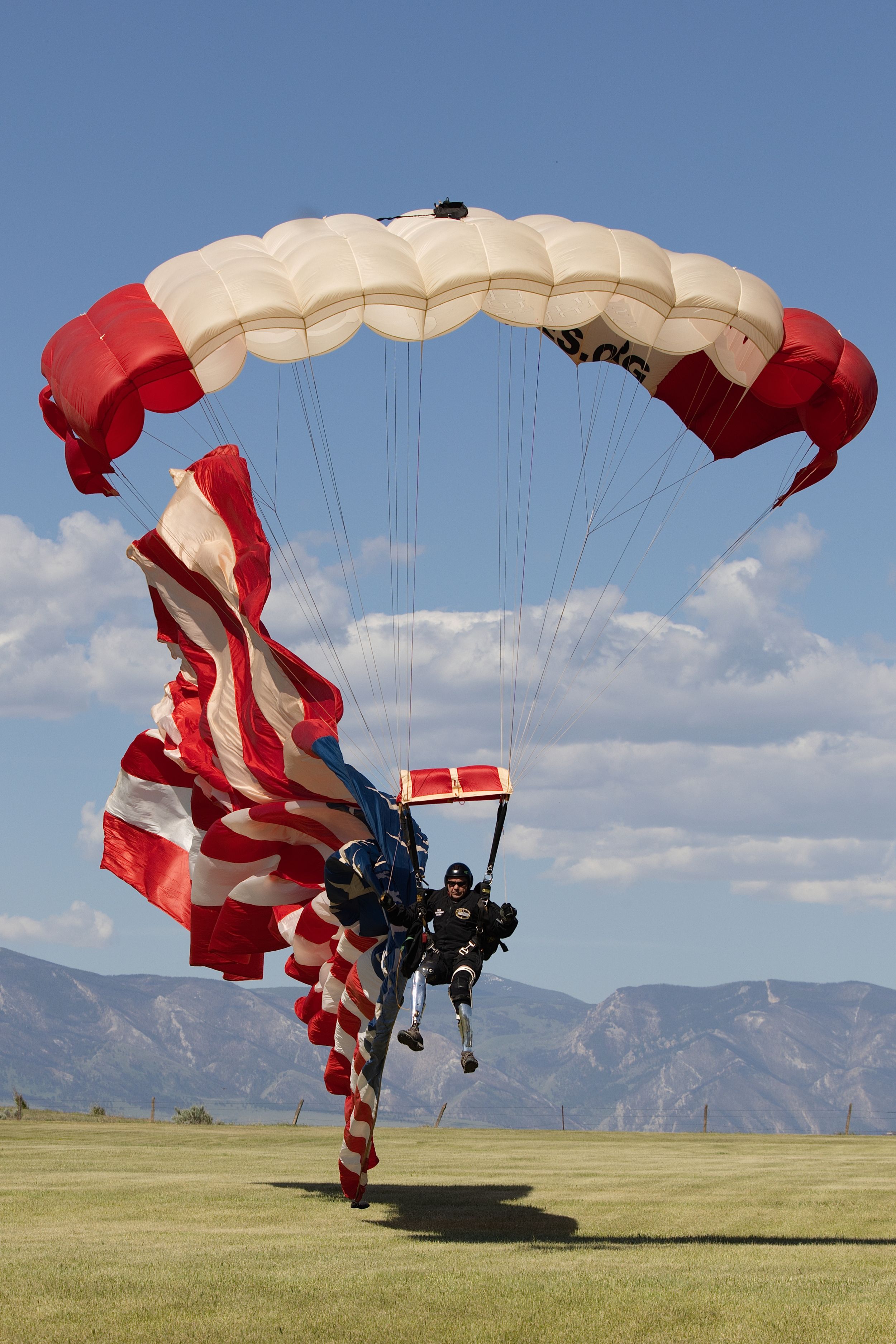 Skydiver landing with American flag under red and white parachute on green field with mountains in background.