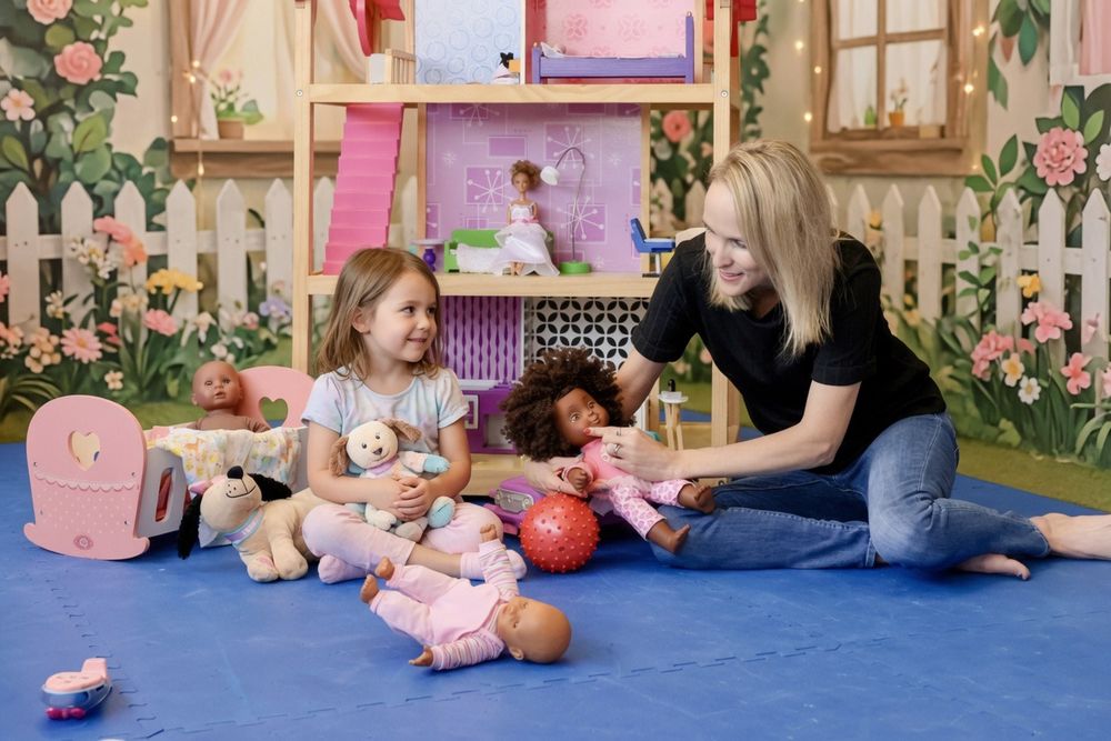 Woman and young girl playing with dolls and toys in a colorful dollhouse playroom setting.