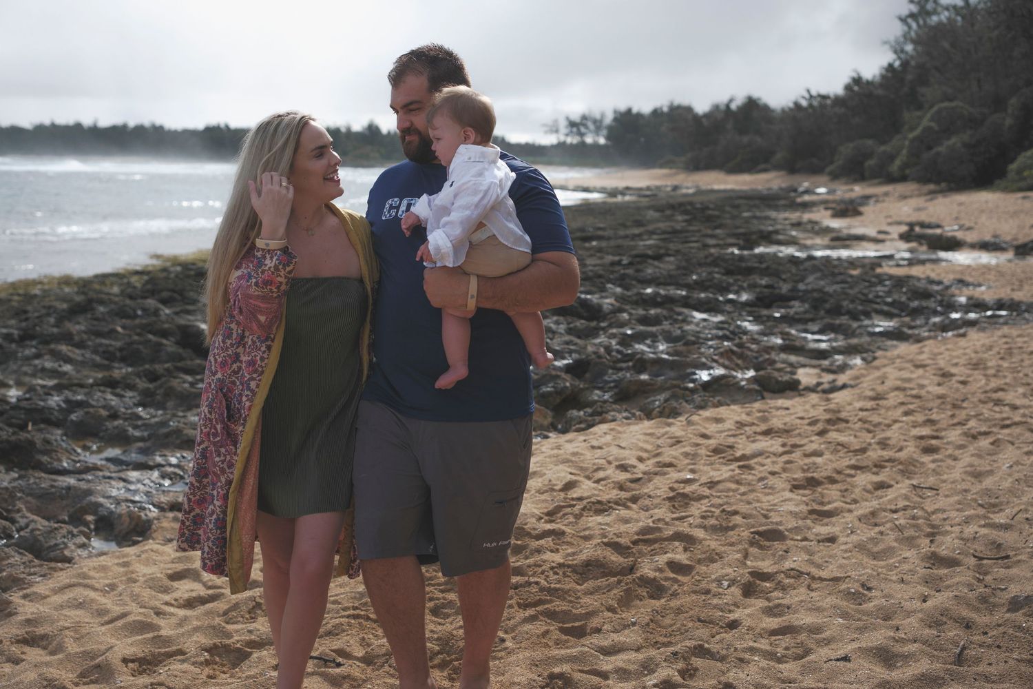Family of three enjoying a beach day, parents smiling at baby on rocky Hawaiian shoreline.