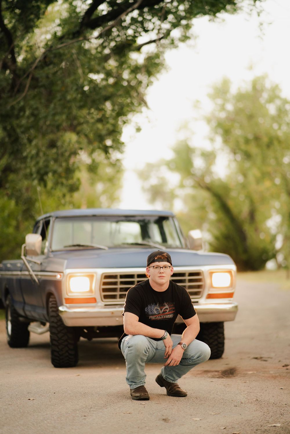 Young man in black t-shirt crouching before a classic Ford pickup truck on a tree-lined dirt road.