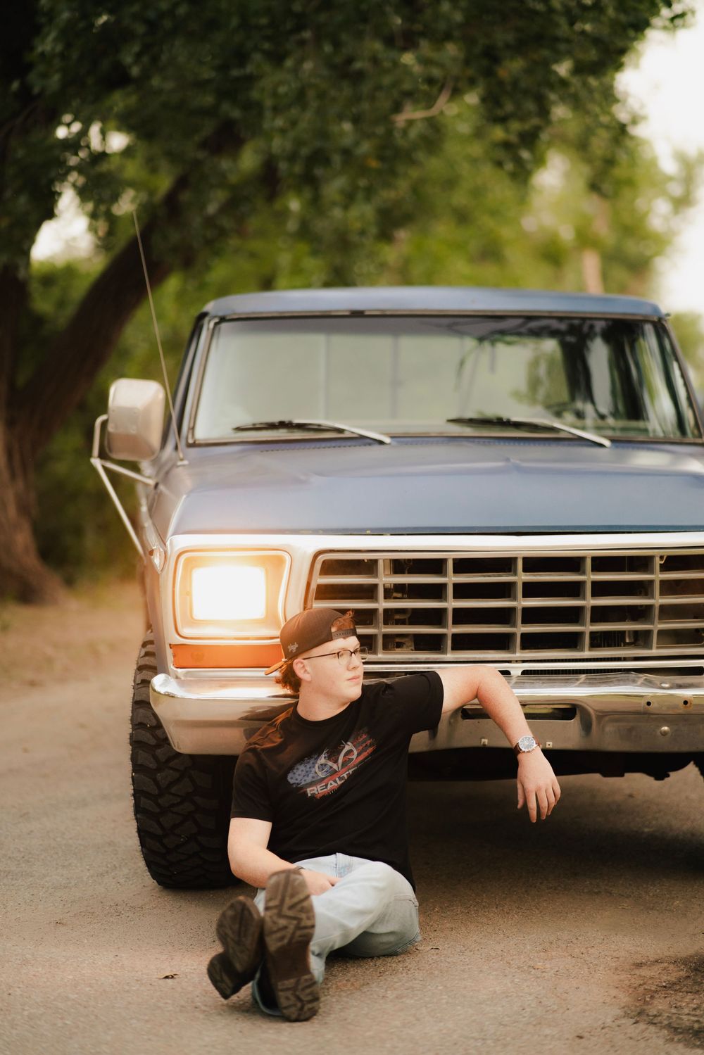 A young person in a black tee sits leaning against a classic blue Ford pickup truck on a dirt road at golden hour.