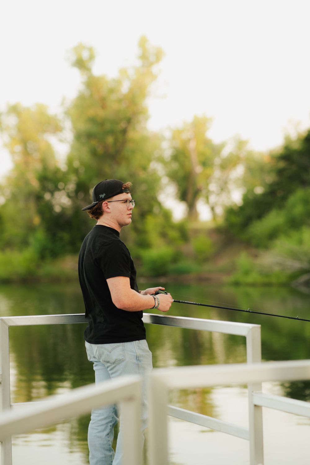 Young man fishing from white dock on calm river, wearing black shirt, cap, and glasses at sunset.