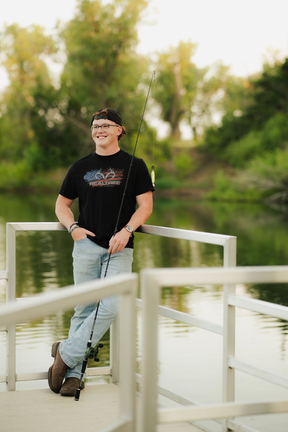 Young man smiling on a fishing dock holding a rod, wearing a black Realtree shirt and jeans near a pond.