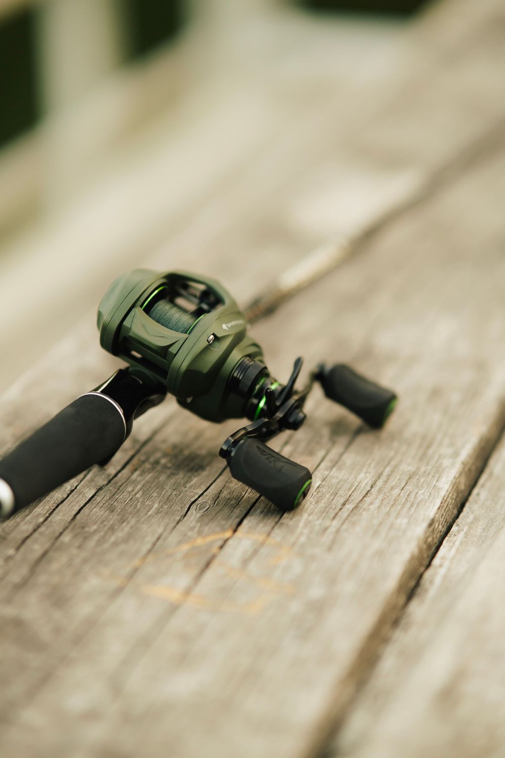 A green baitcasting fishing reel mounted on a rod, resting on a weathered wooden dock surface.