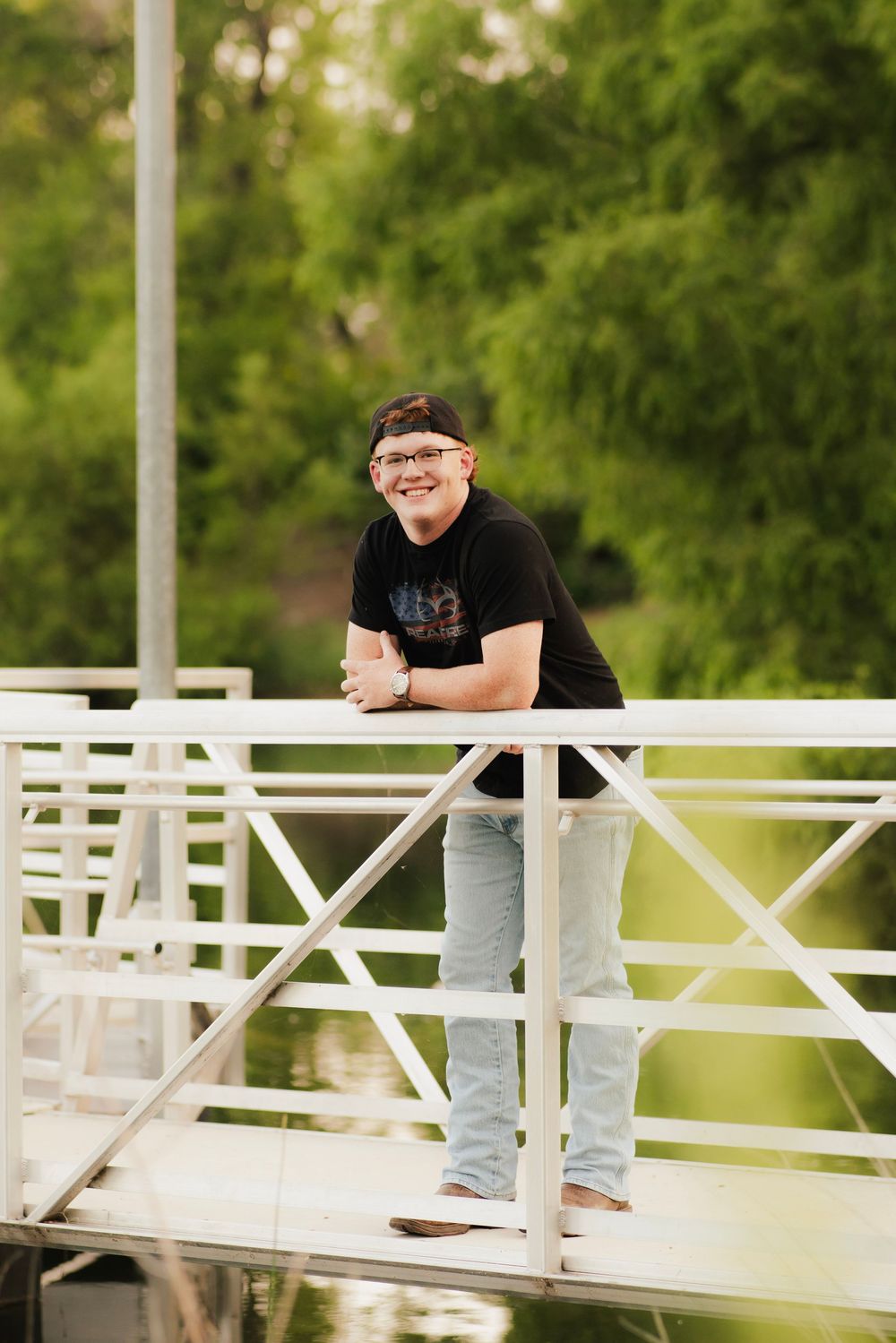 Young person in black t-shirt and jeans smiling on a white dock bridge surrounded by lush green trees.
