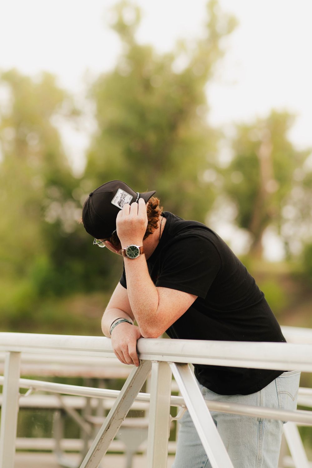 Person in black t-shirt and cap leaning on white railing, covering face with hand, wearing a watch outdoors.
