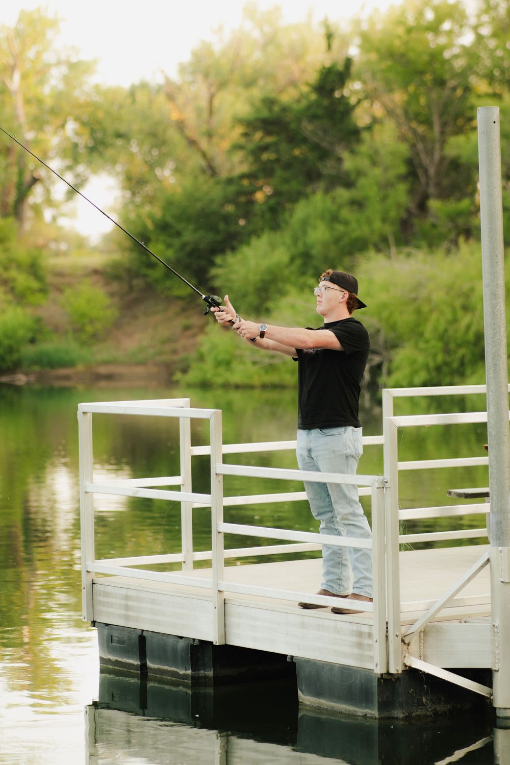 Young man in black shirt and jeans fishing from a white dock surrounded by lush green trees and calm water.