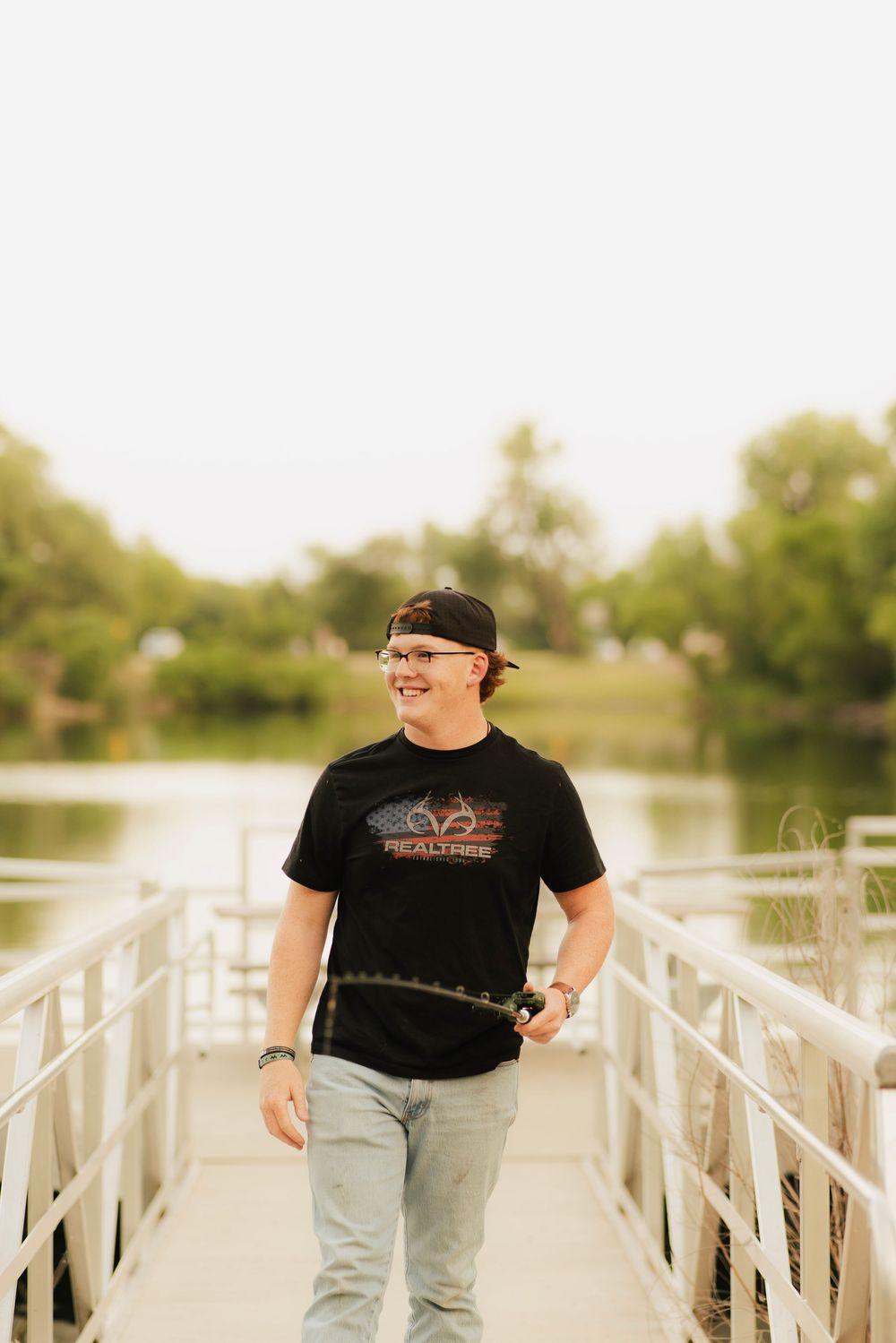 Smiling teen in Realtree shirt and cap walks on a dock by a calm lake surrounded by green trees.