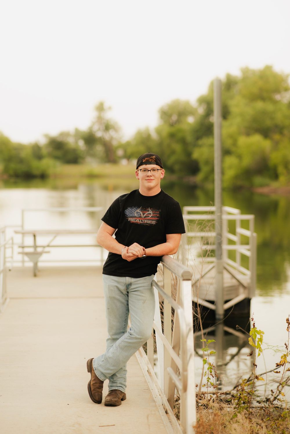 Young man in black graphic tee and jeans leaning on a dock railing by a calm river surrounded by lush green trees.