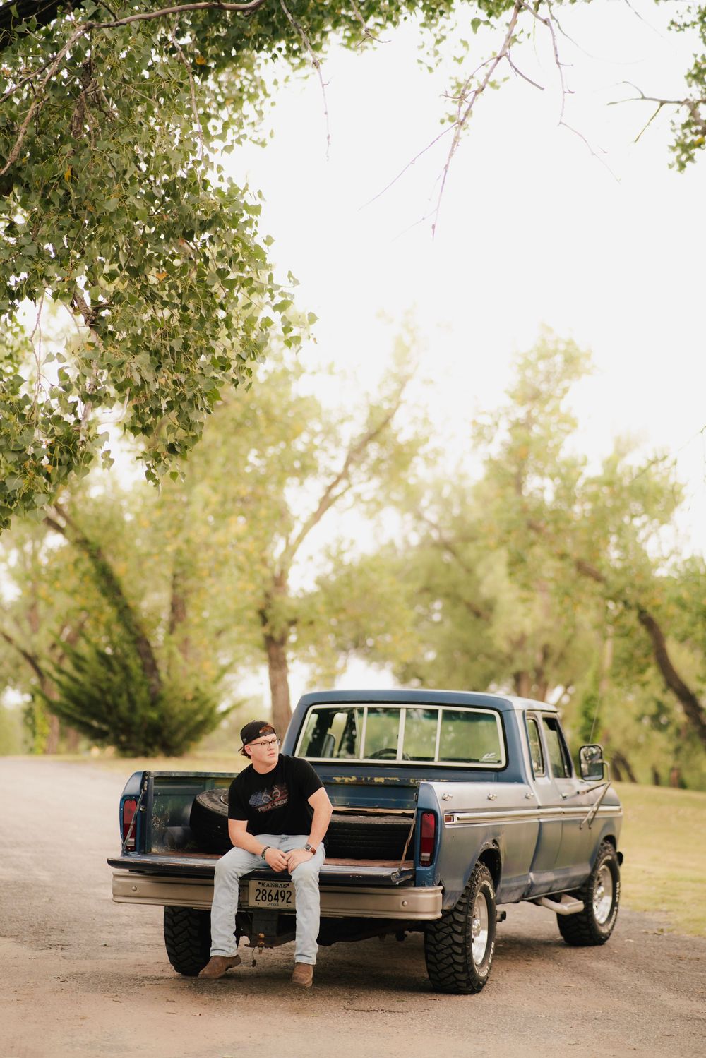 Young man sitting on the tailgate of a classic Ford pickup truck on a rural dirt road surrounded by trees.
