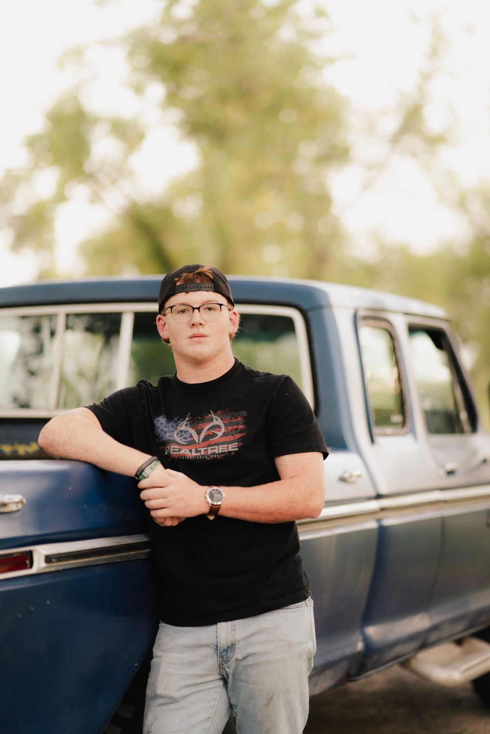 Teen in Realtree shirt leans against vintage blue truck outdoors at golden hour.