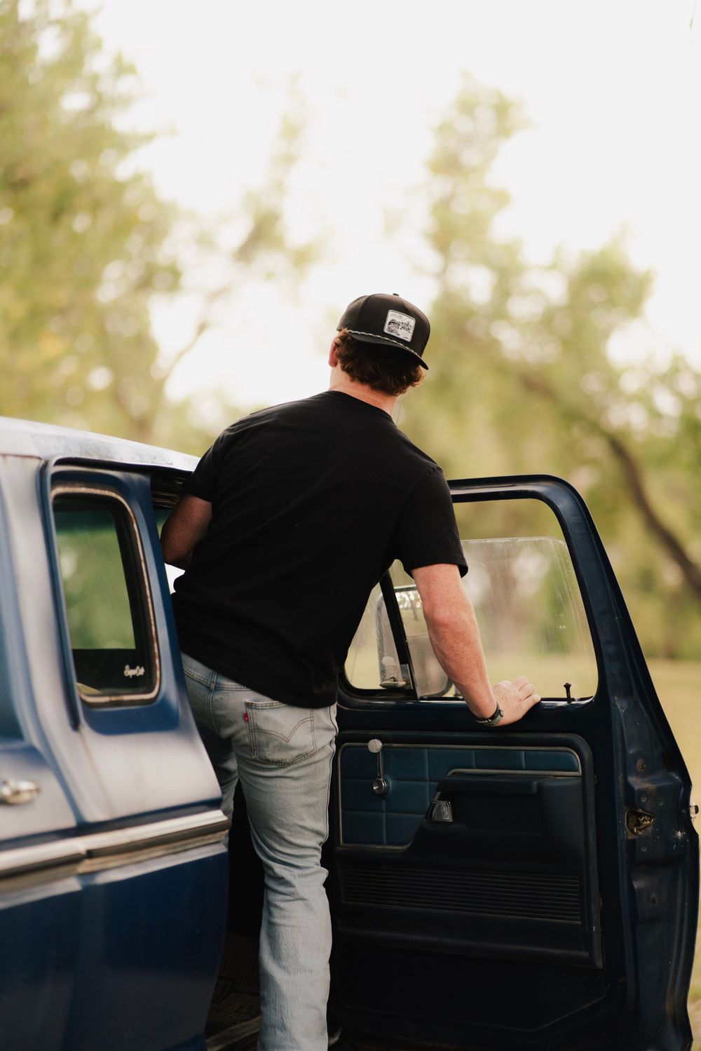 Man in black t-shirt and cap opening door of vintage blue truck outdoors.