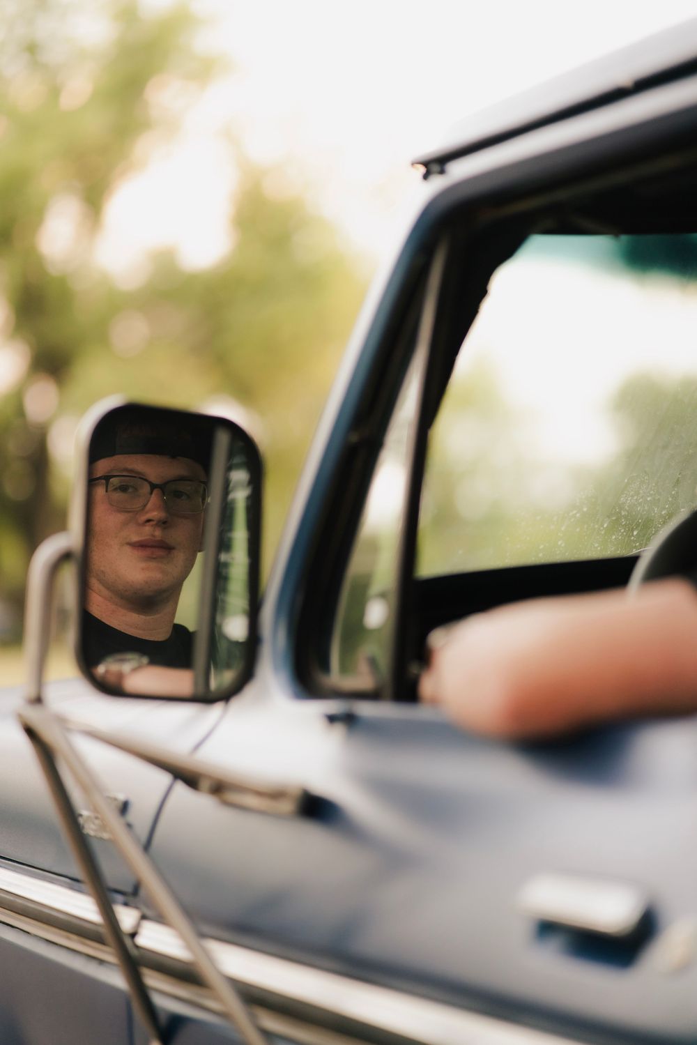 Teen in glasses and camo cap reflected in vintage truck side mirror outdoors.