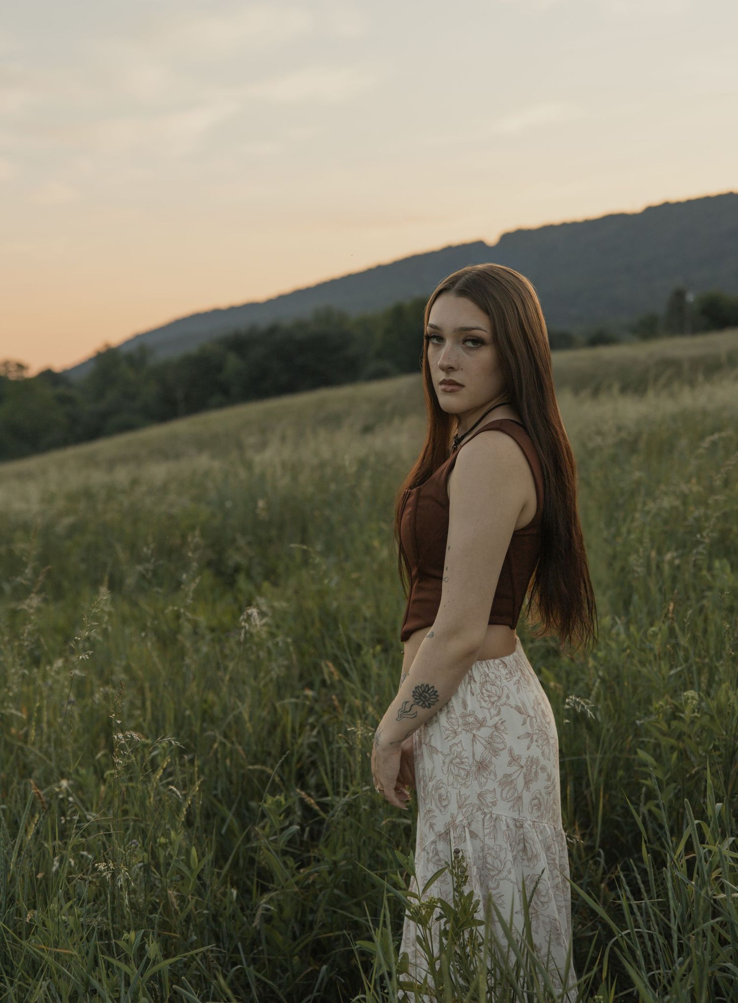 Young woman with long red hair in brown crop top and floral skirt posing in meadow at sunset.