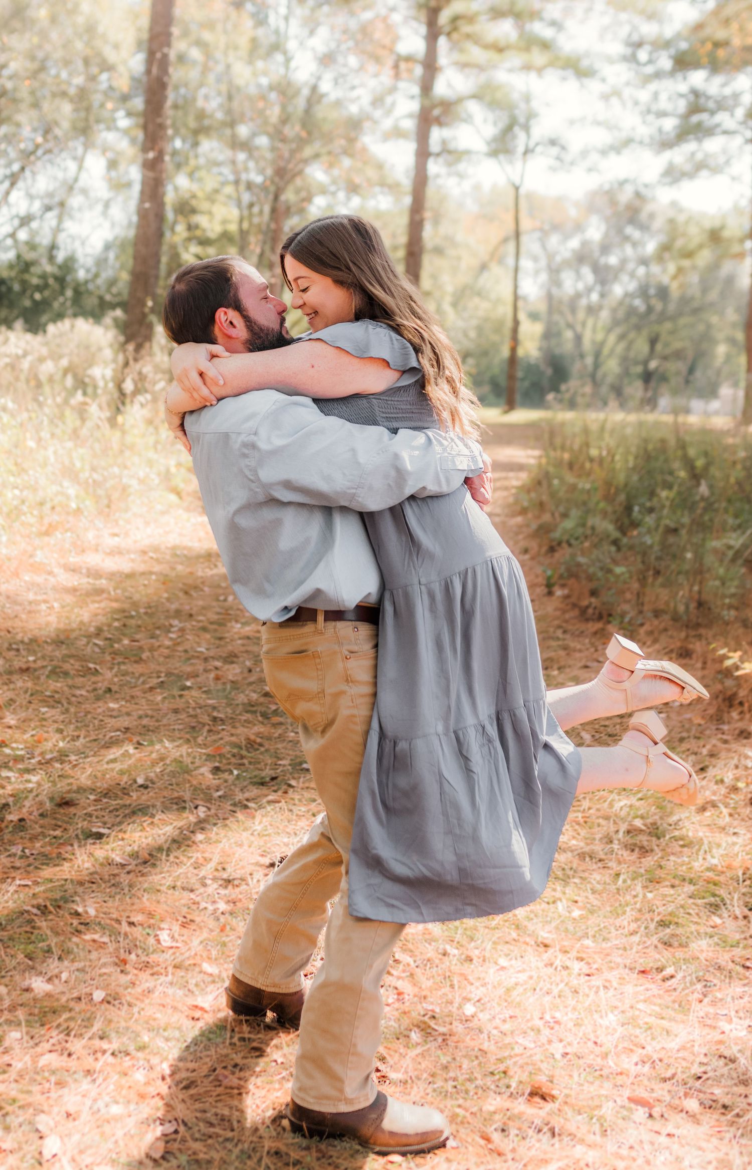 A man lifts and embraces a woman in a gray dress outdoors, sharing a romantic moment on a sunny woodland path.