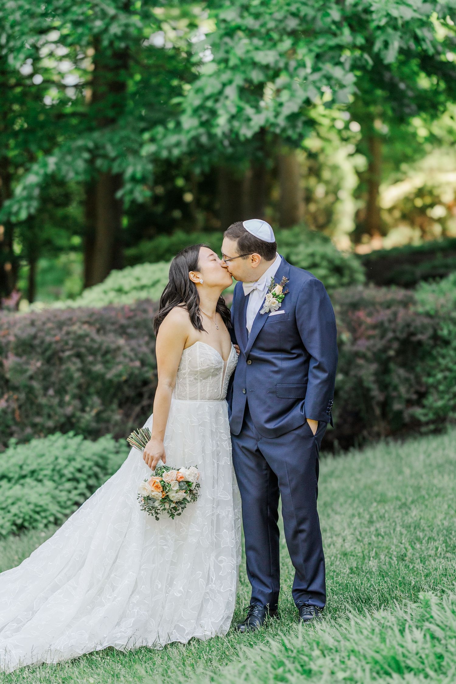 A bride and groom share a kiss outdoors, she holds a floral bouquet in a lace gown, he wears a navy suit and kippah.