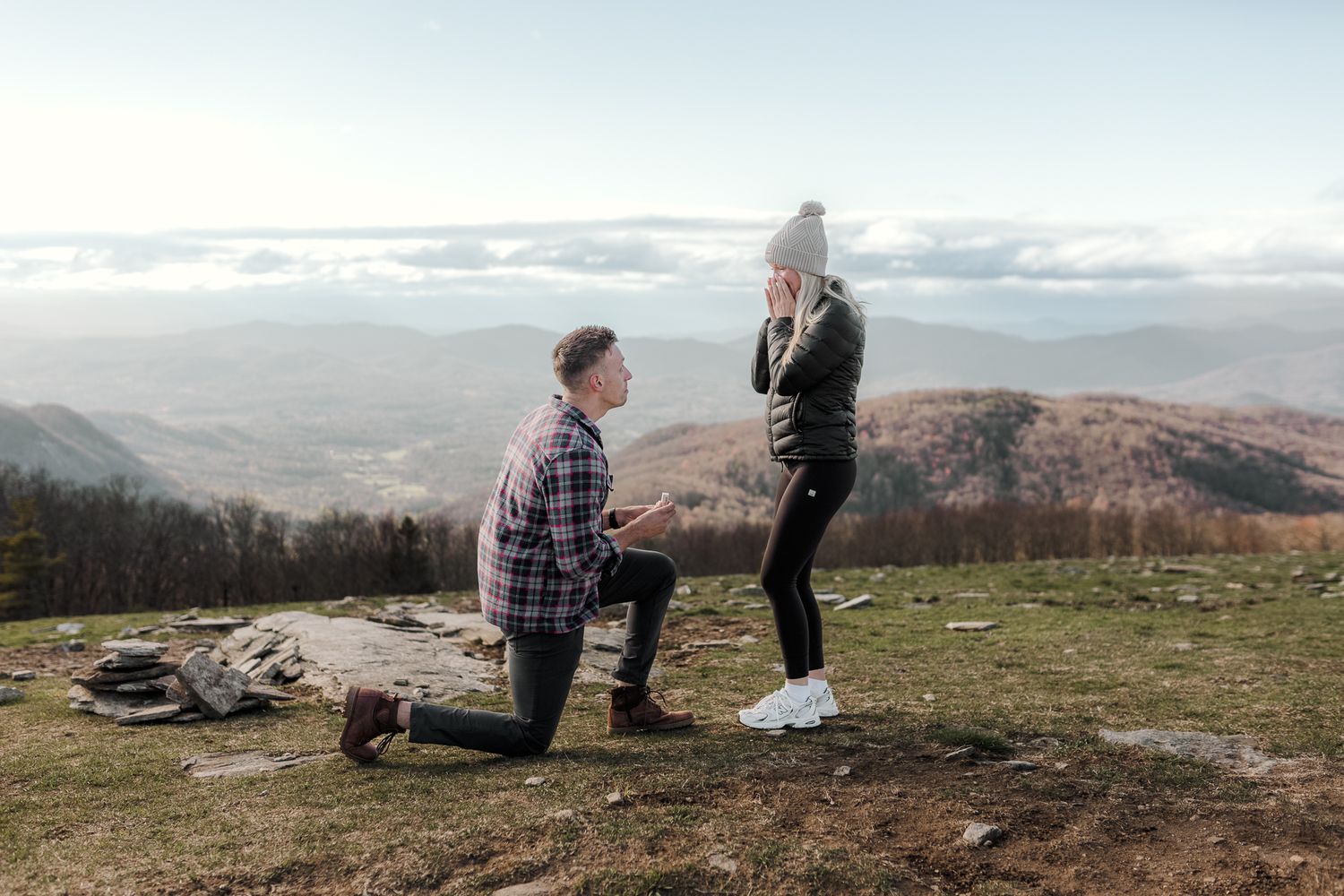 Man proposing on one knee to woman on mountain overlook with scenic valley and clouds below.