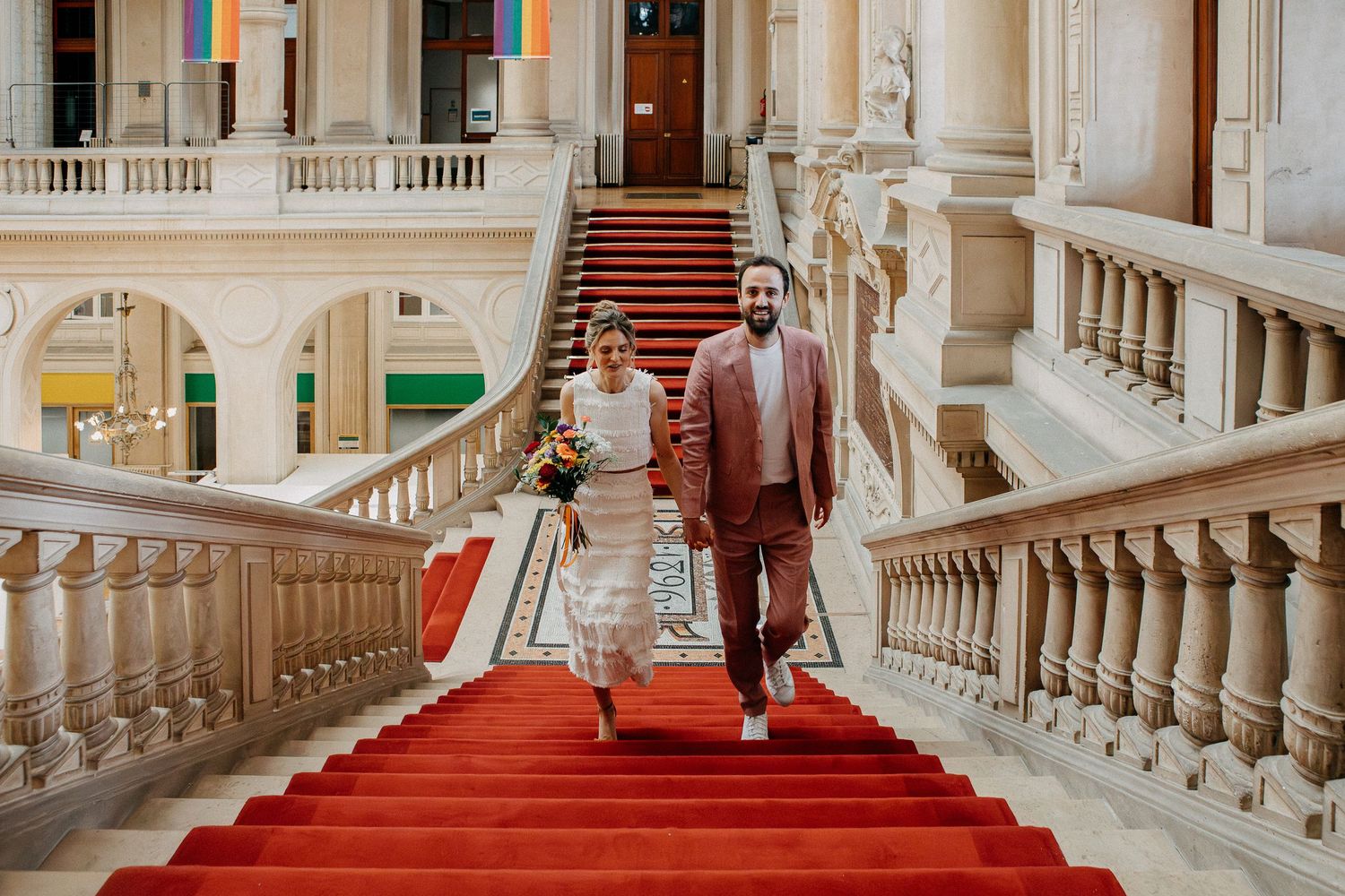 Bride and groom descend grand staircase with red carpet in elegant wedding venue.