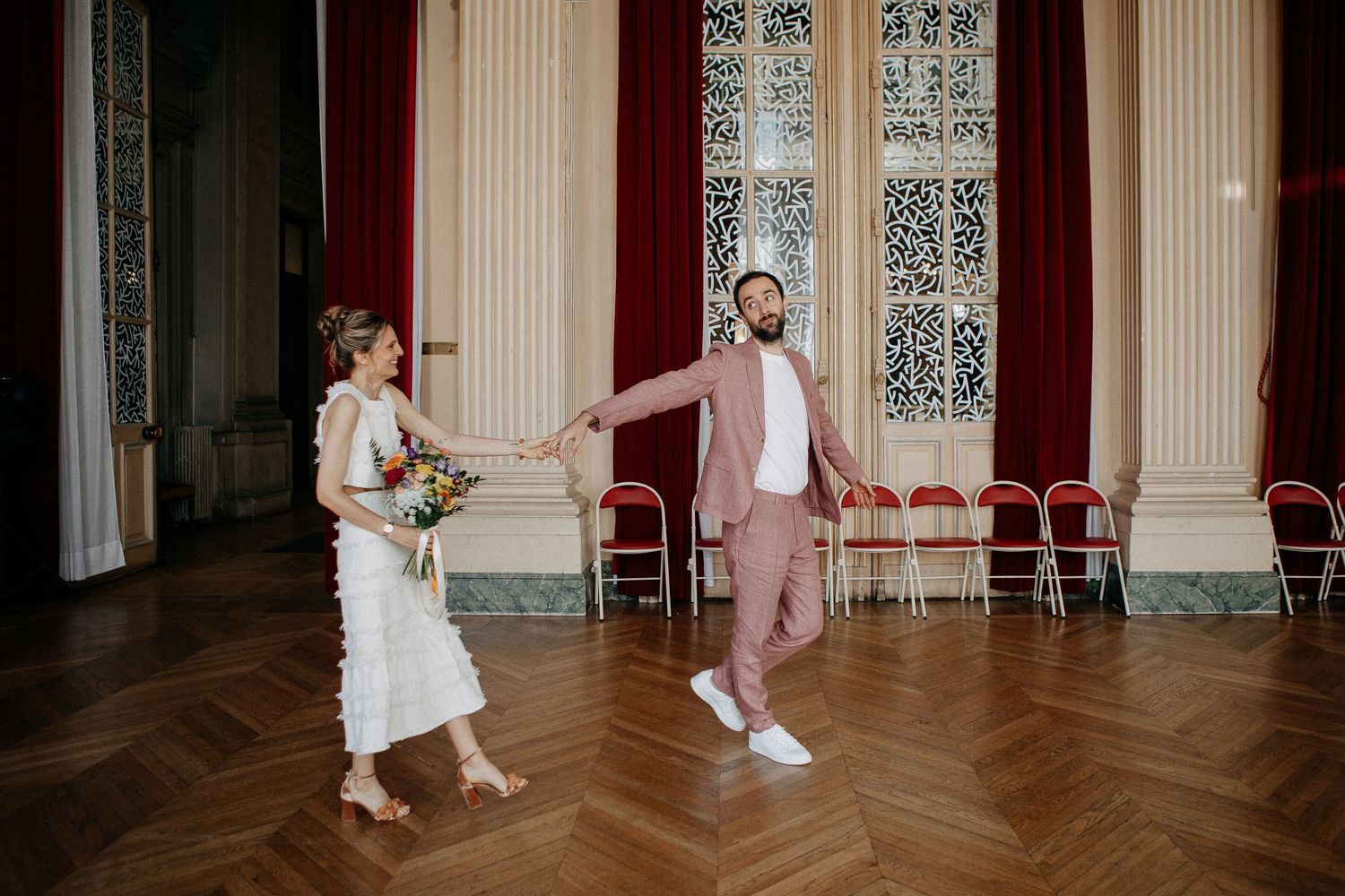 Bride and groom dancing joyfully in an elegant ballroom with parquet floors and tall windows.