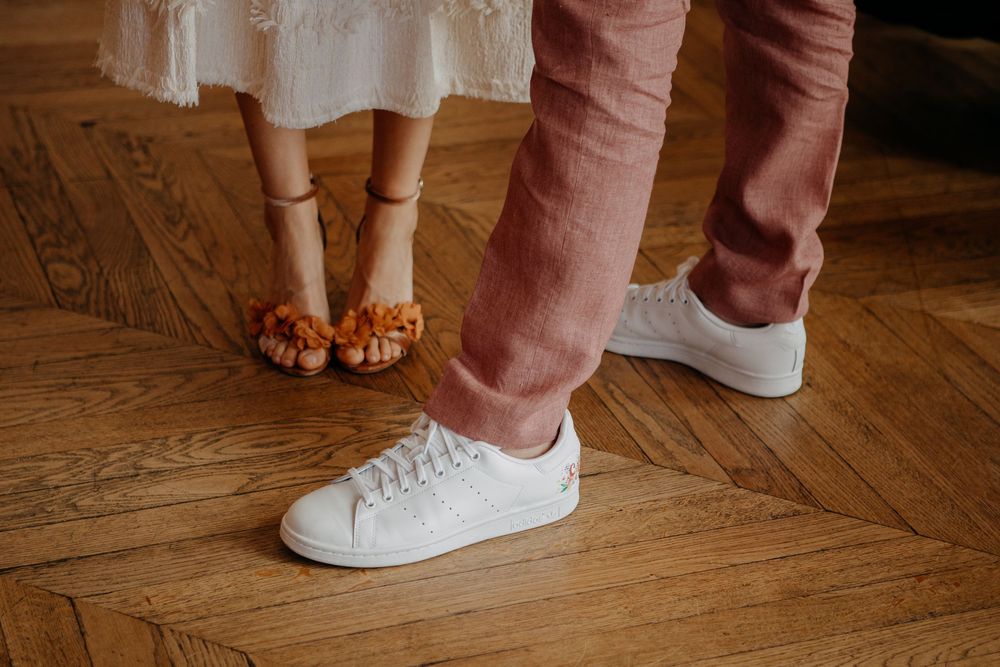 Close-up of a couple's feet, woman wearing orange fringe sandals and man in white sneakers, standing on herringbone wood floor.