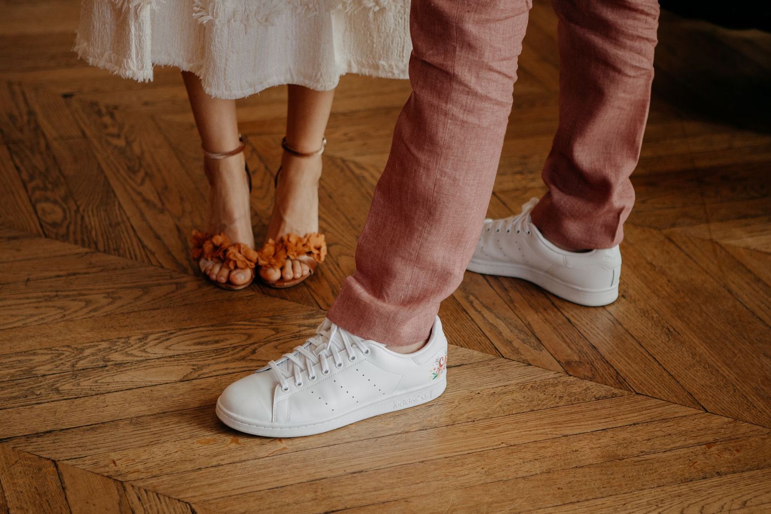 Close-up of a couple's feet, woman wearing orange fringe sandals and man in white sneakers, standing on herringbone wood floor.