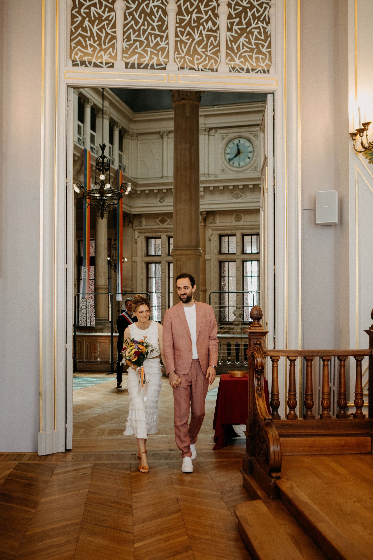 Couple walking hand-in-hand through elegant venue doorway, groom in pink suit, bride holding bouquet in white dress.