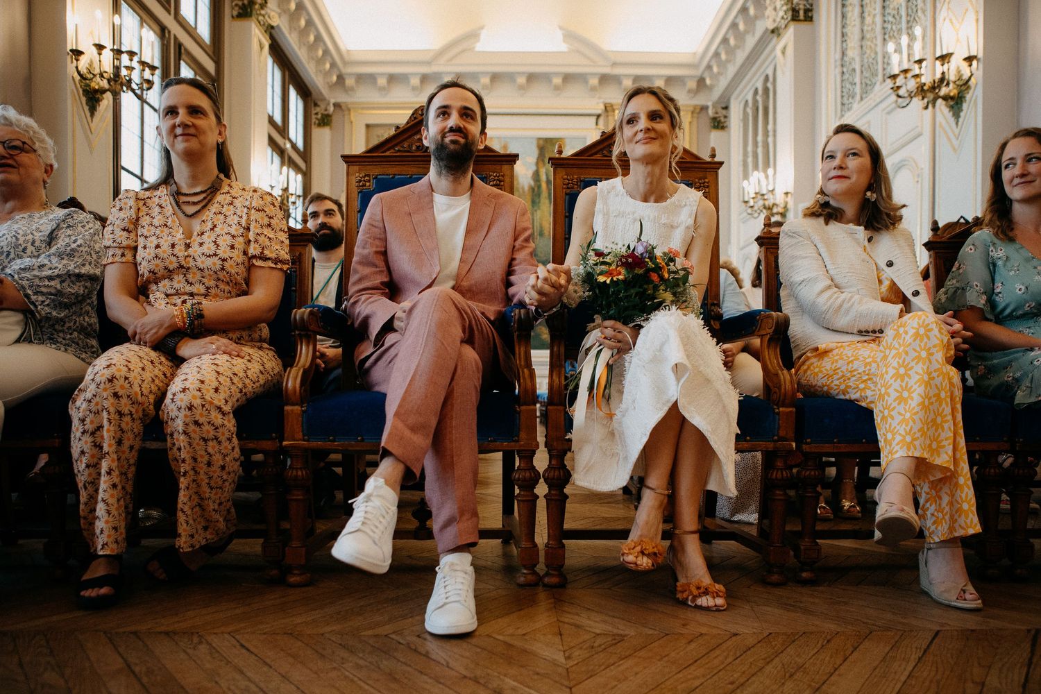 Bride and groom seated at civil wedding ceremony with guests, bride holding bouquet, groom in pink suit.