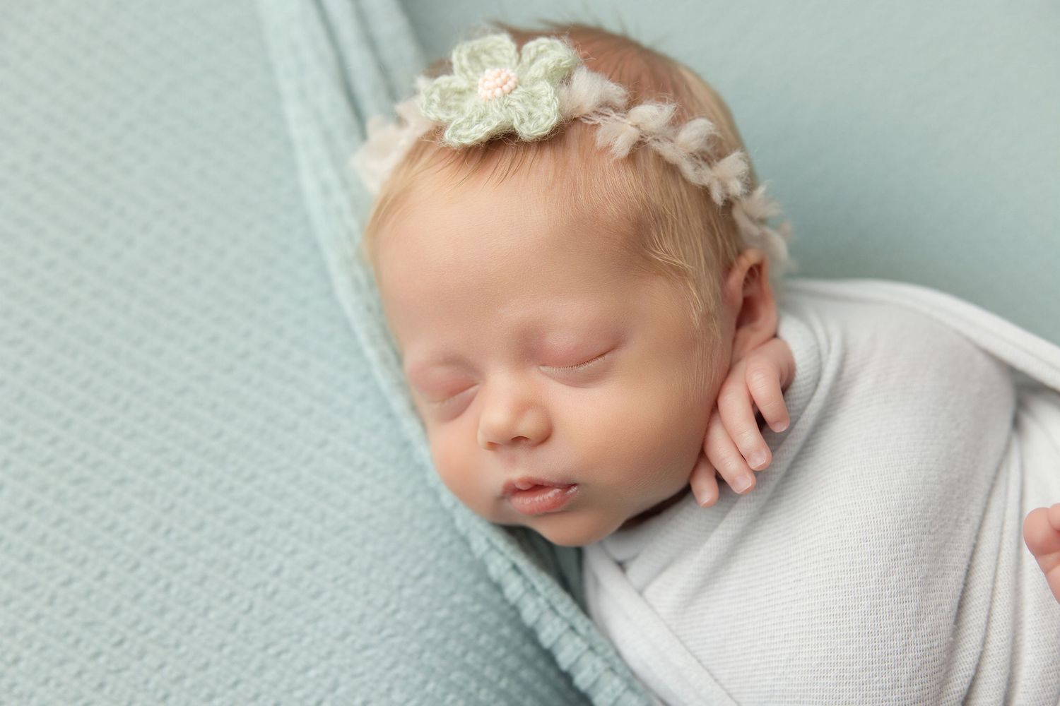 Newborn baby in teal hooded knit onesie with wooden buttons, sitting in a vintage metal bucket with soft teal blankets.