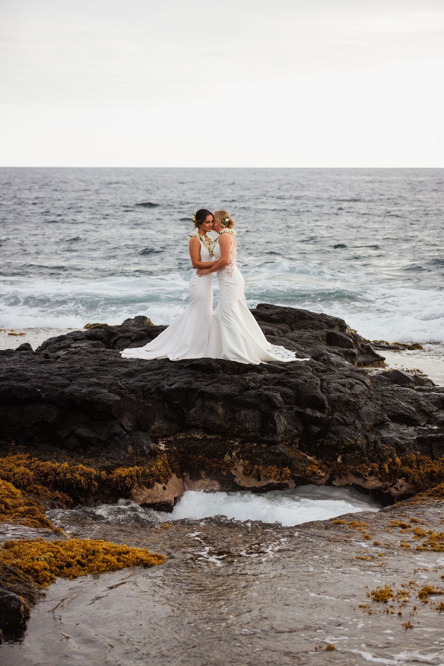 Two brides in white mermaid gowns embrace on rocky ocean shore with waves in background.