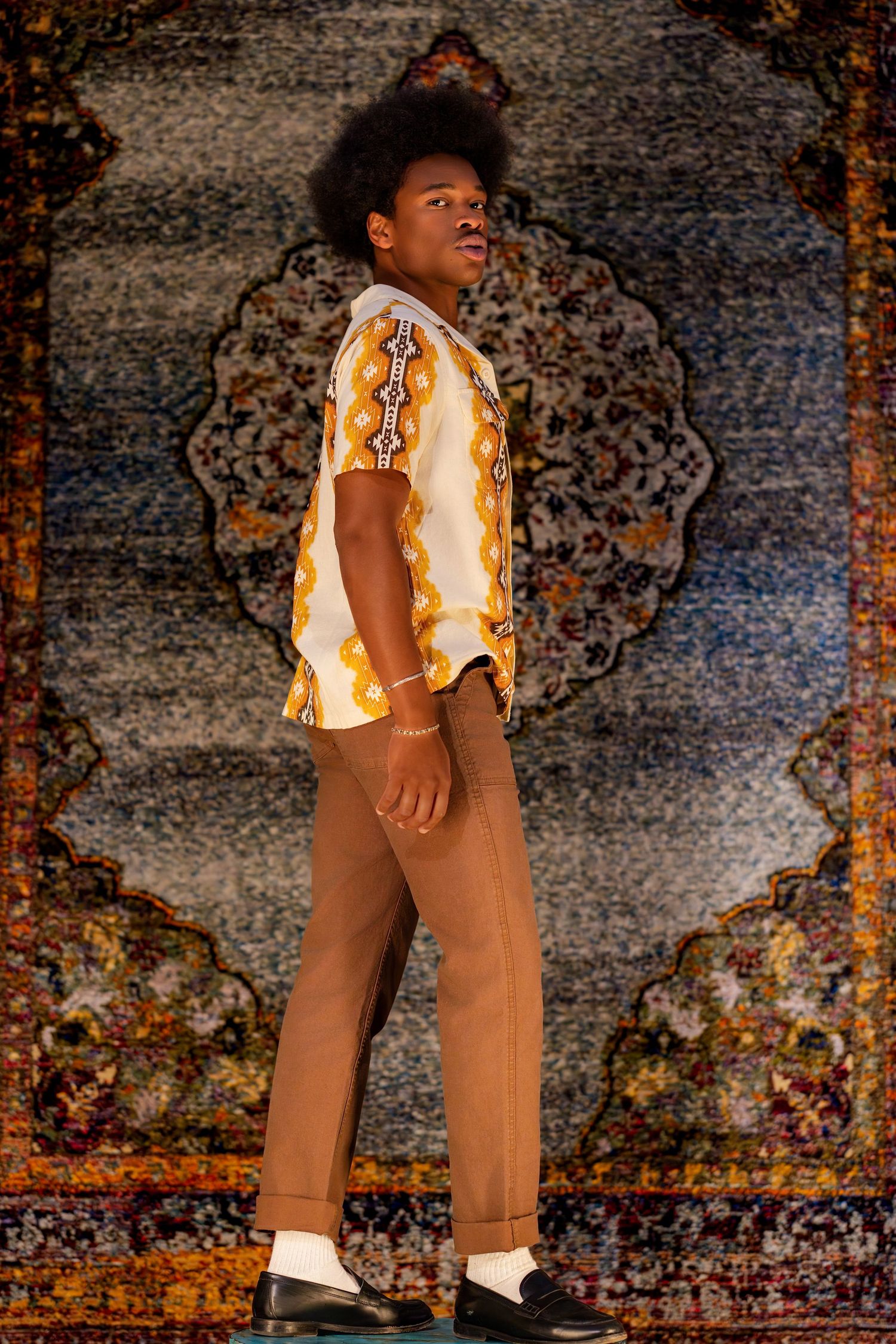 Young Black male model poses in patterned shirt and tan trousers before ornate tapestry.