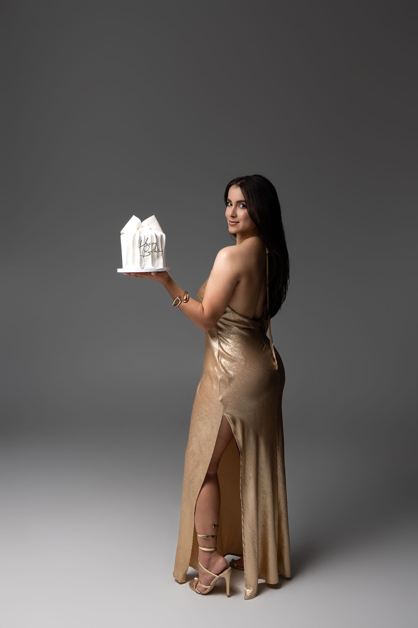Woman in gold gown holding white architectural model, posing elegantly against gray background.