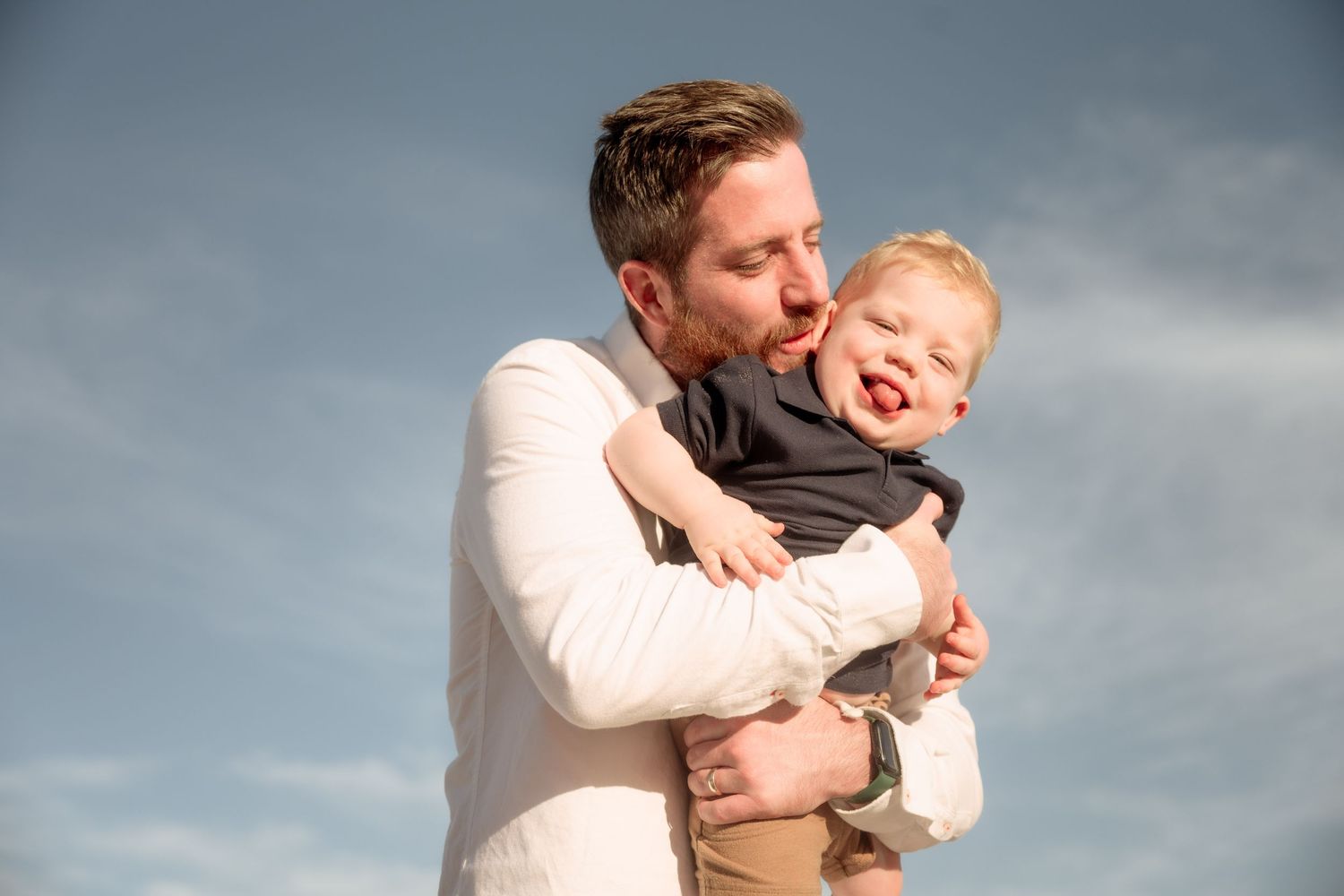 A father lovingly holds his laughing toddler against a blue sky backdrop.