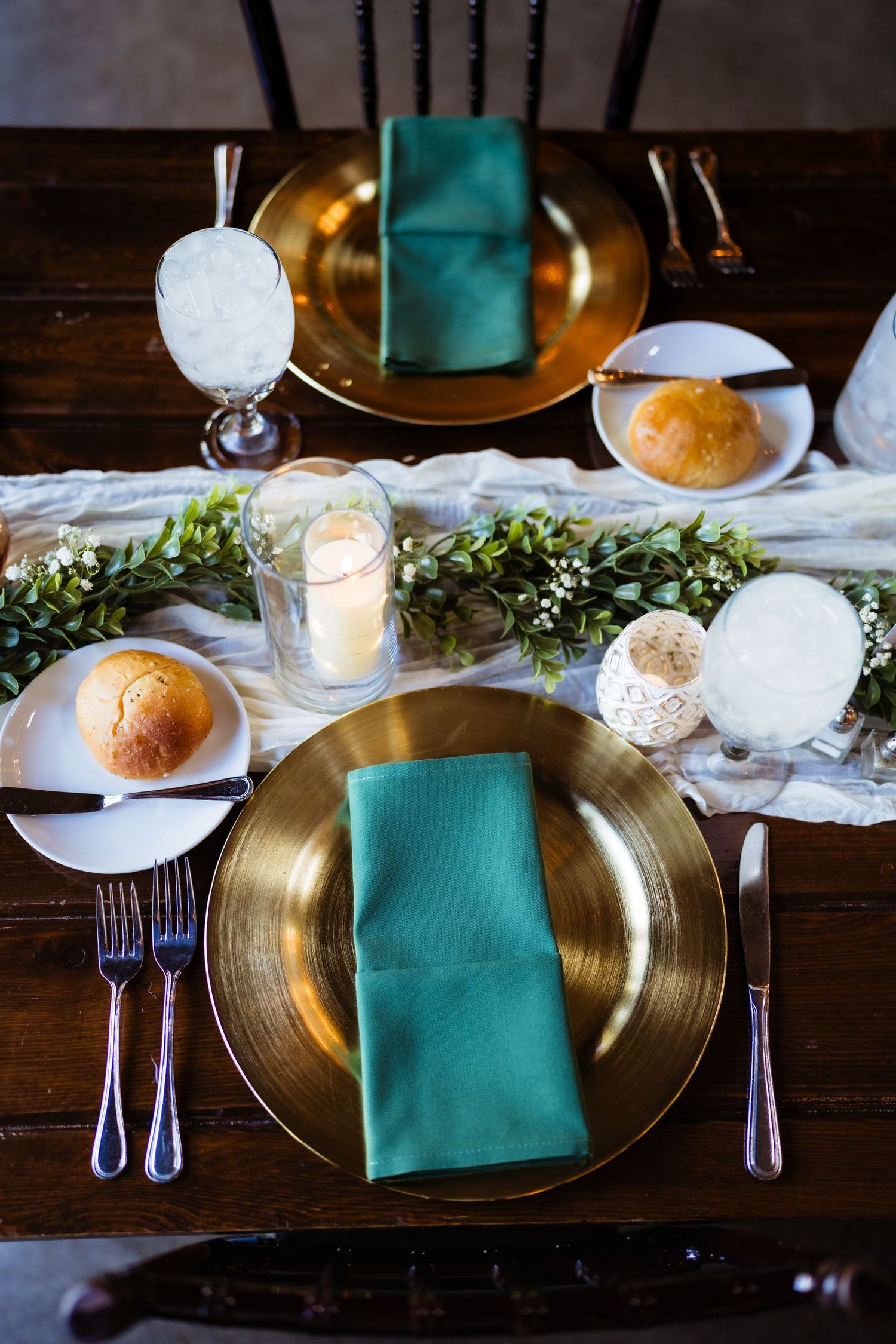 Elegant table setting with gold charger plates, teal green napkins, candles, greenery runner, and fresh bread rolls on dark wood table.