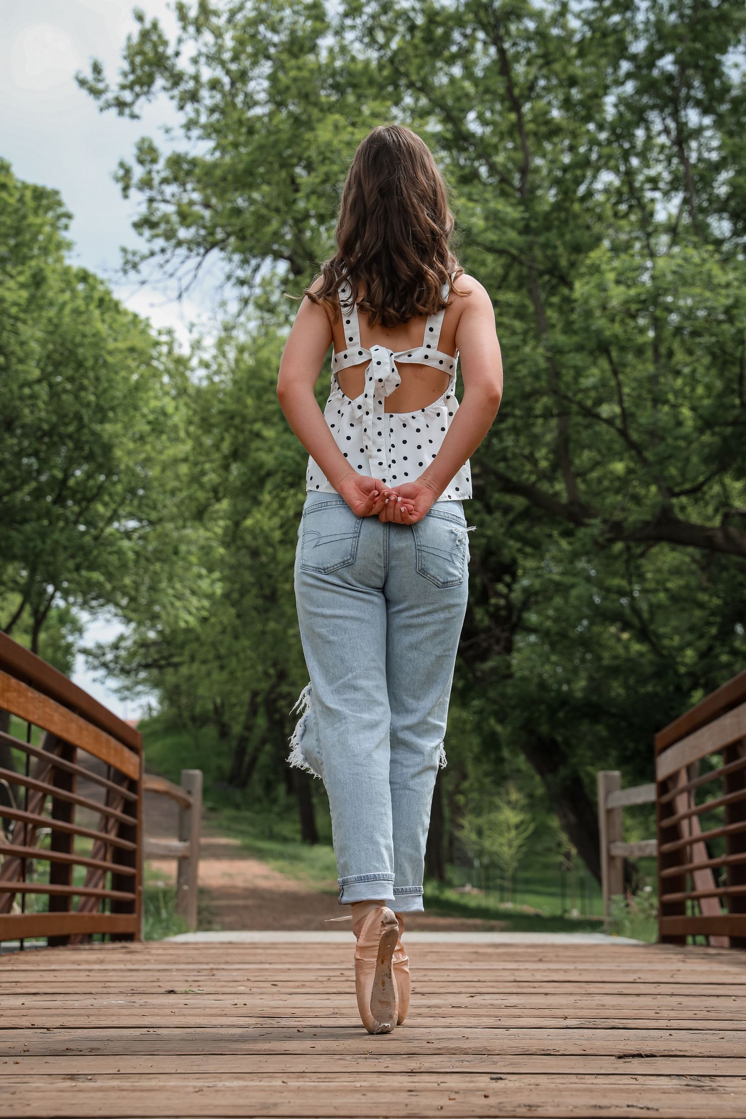 Young woman in jeans and polka dot top standing on tiptoe on a wooden bridge surrounded by green trees.