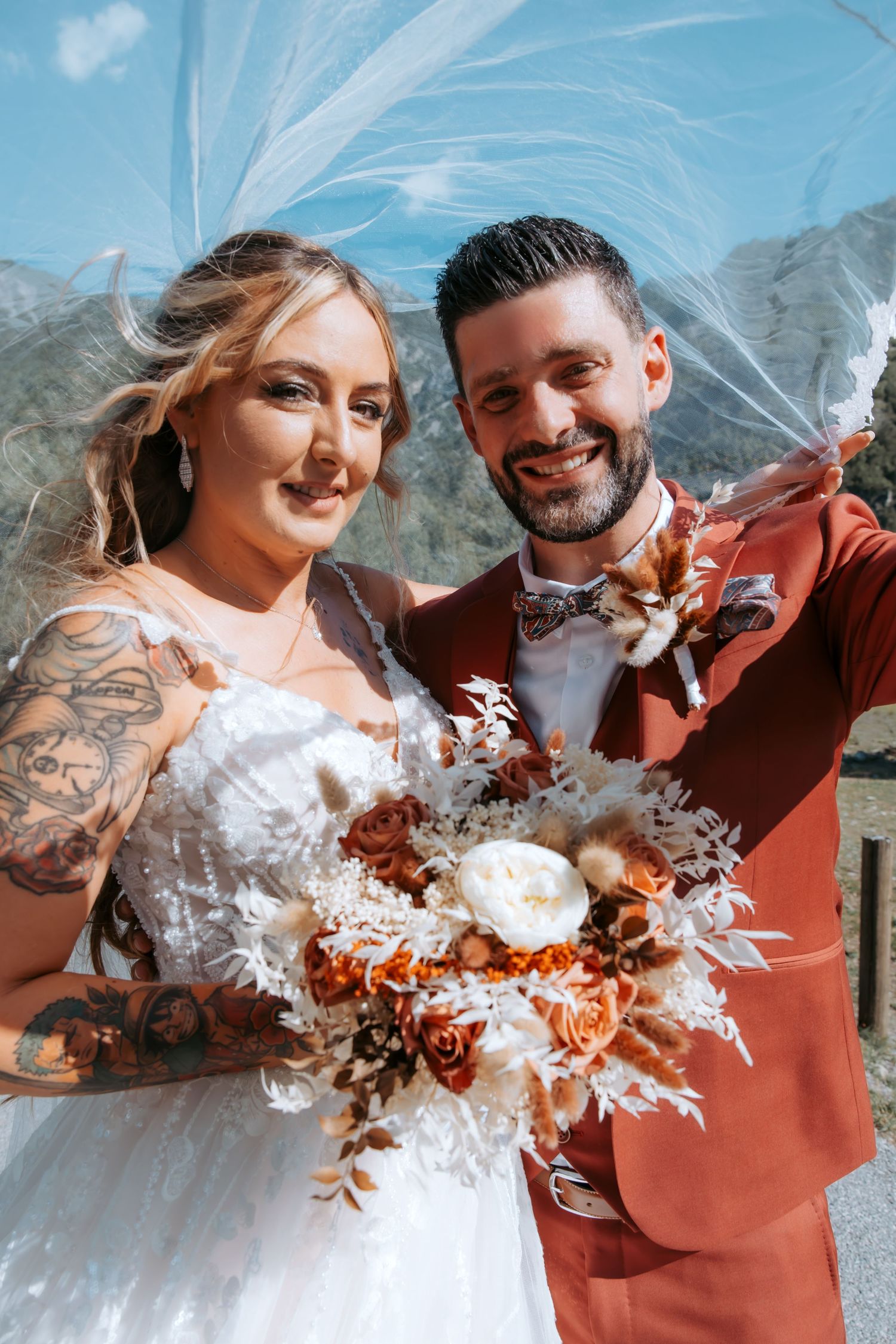 Bride with tattoos in white dress holding autumn bouquet stands with groom in rust suit under veil with mountains behind.