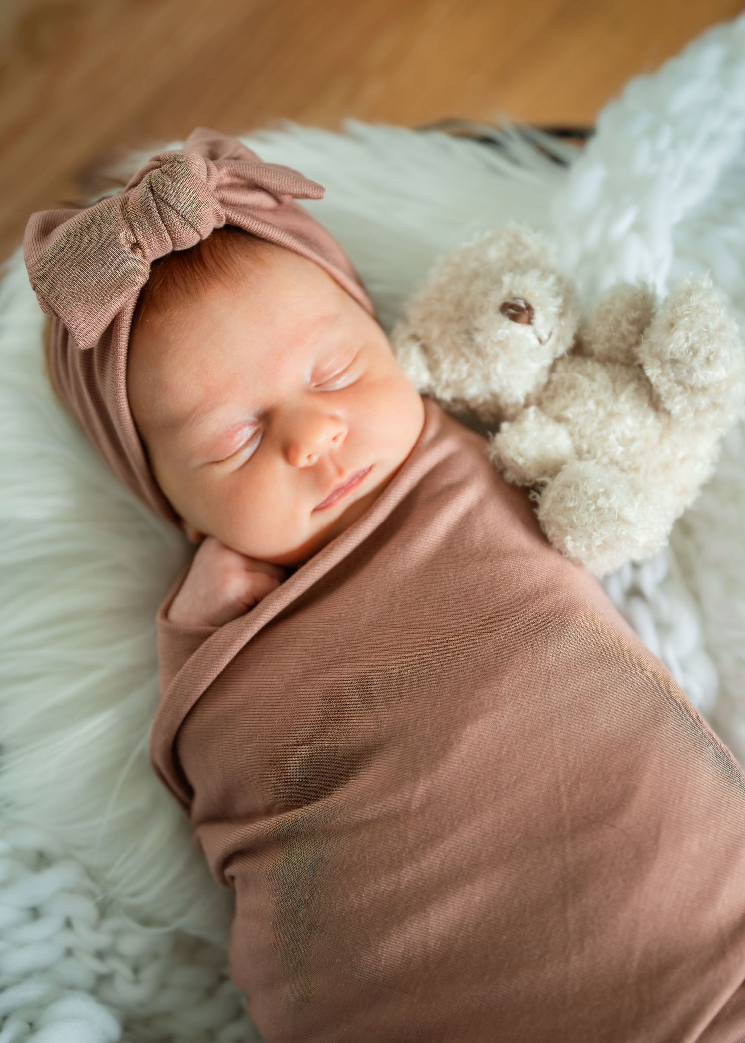 Sleeping newborn baby swaddled in mauve wrap with bow headband beside a soft white teddy bear on fluffy blanket.