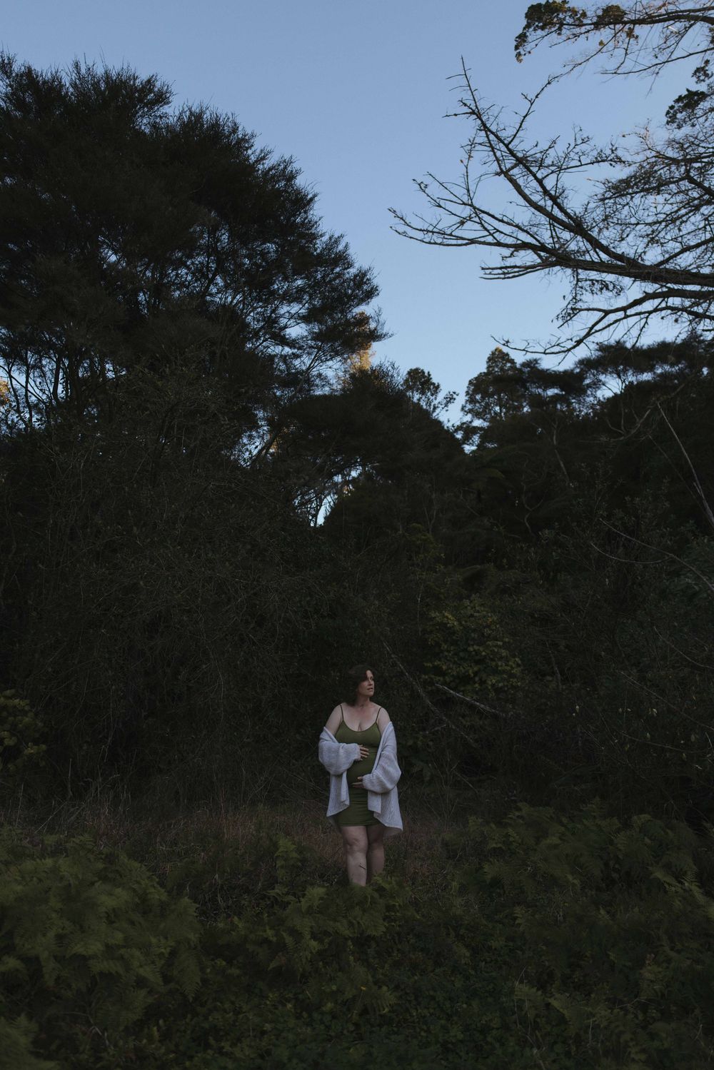 Woman in white wrap standing among lush dark forest greenery at dusk with tall trees in background.