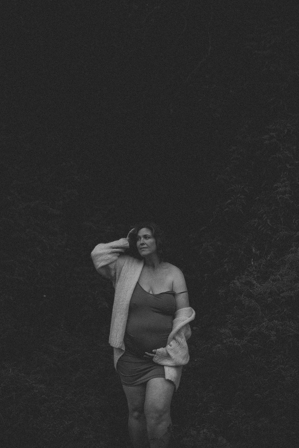 Black and white maternity photo of a pregnant woman in a bodysuit and cardigan standing outdoors among dark trees.
