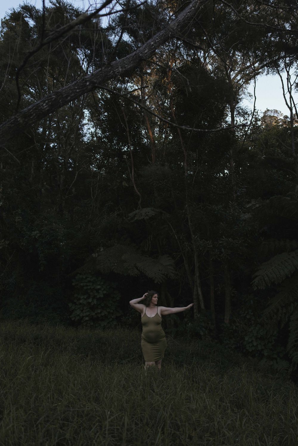 Woman in olive dress stands with arms outstretched in a dark, lush forest clearing at dusk.