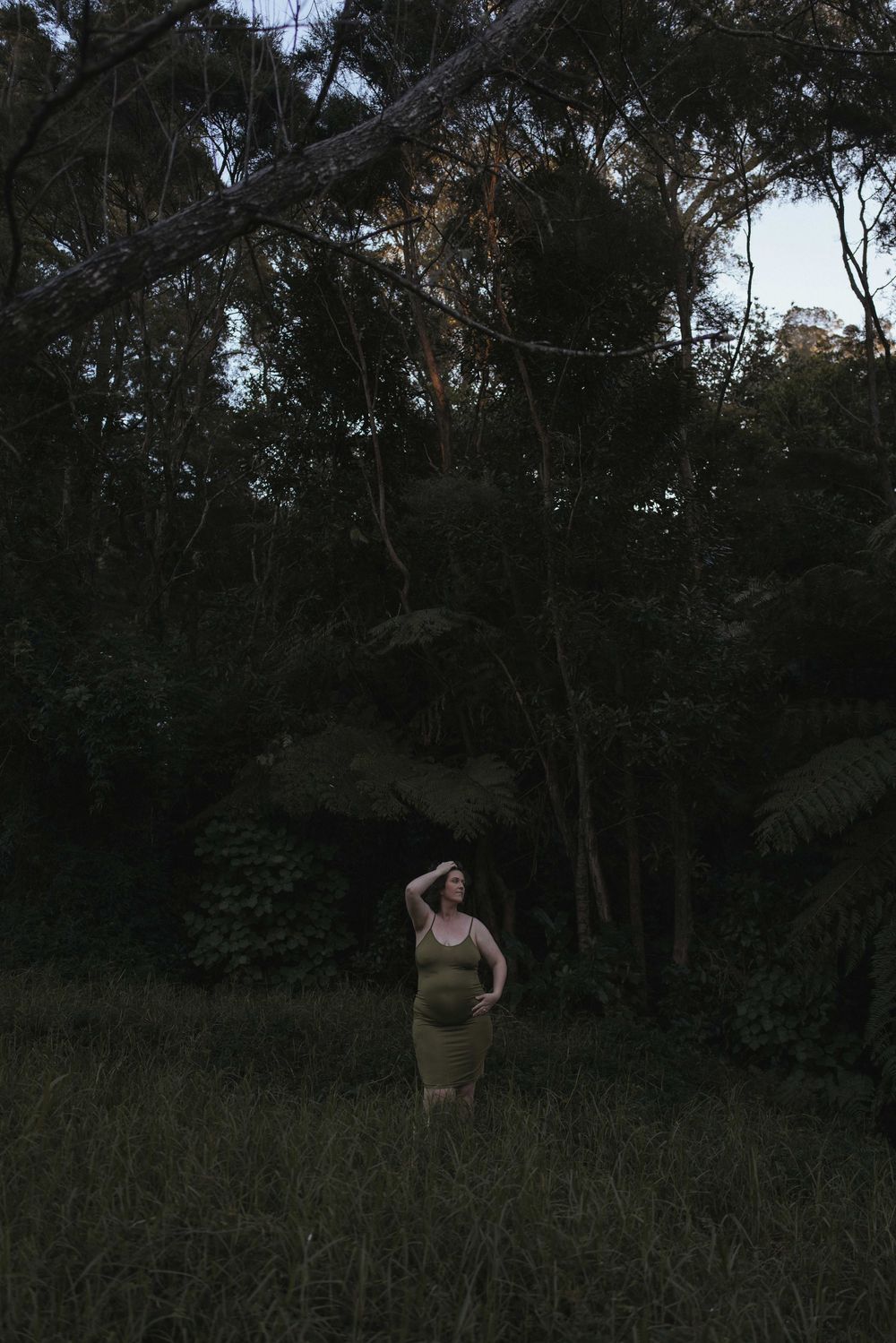 Pregnant woman in olive dress stands in dark woodland clearing at dusk, hand raised to forehead.