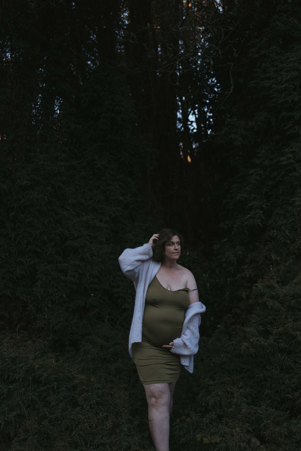 Pregnant woman in olive dress and white cardigan standing among dark trees at dusk, cradling her baby bump.