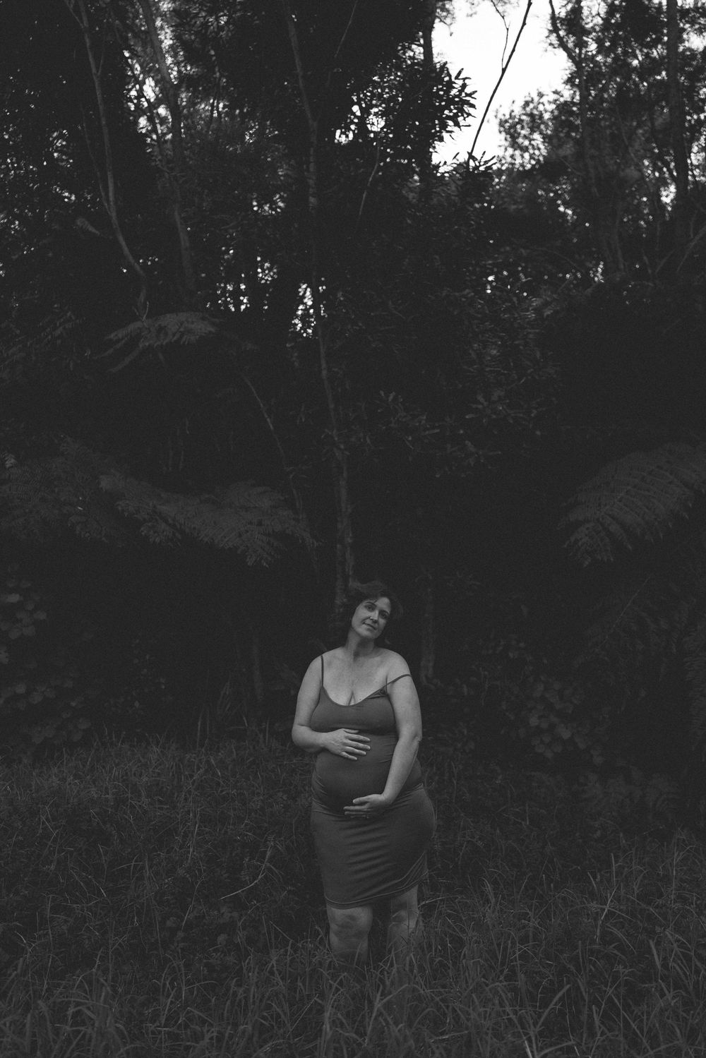 Black and white photo of pregnant woman in form-fitting dress cradling baby bump in lush forest setting.